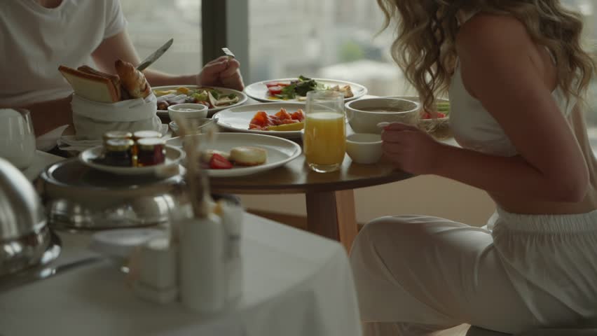 A cinematic shot with unrecognizable faces. A young couple, a man and a woman in white clothes, are sitting having breakfast in a hotel on the top floor of a skyscraper. Healthy meals at the hotel Royalty-Free Stock Footage #3400438657