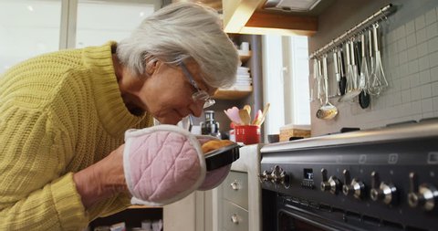 Happy old Caucasian lady wearing oven gloves taking out freshly baked muffins from oven 4K 4k