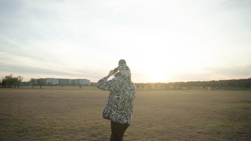Wide shot of dad running through grassy field at sunset with his baby on his shoulders. Slow motion