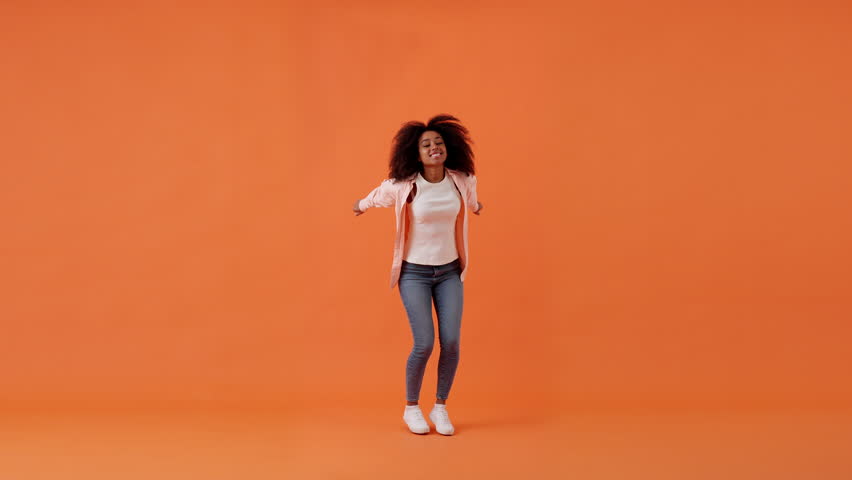 Cheerful African American woman jumps up enjoying moment of life in studio with orange background. Positive emotions and shooting in modern photo studio slow motion