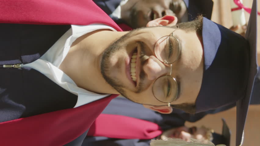 Vertical portrait of positive graduate in hat and gown looking at camera while his collegemates celebrating in background