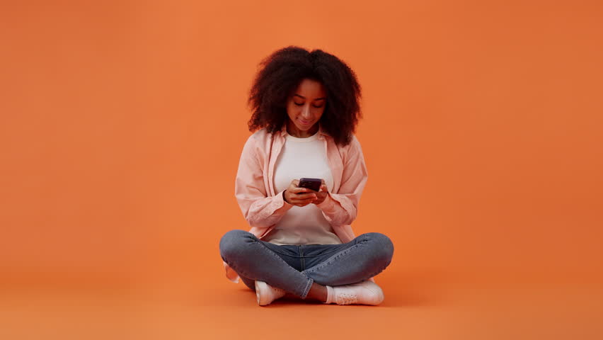 Happy African American woman corresponds with partner on mobile phone thinking about content of message sitting on floor in orange studio. Maintaining long-distance relationship Royalty-Free Stock Footage #3400501415
