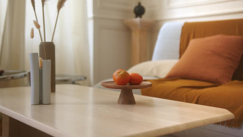 Slow zoom in shot of a living room table with some furnishing objects on. On the background there is a sofa, a pillow and a blanket. Clear colors and boiserie on the walls. Bright room