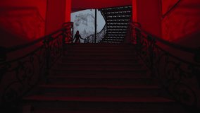 In a wide view, a gothic woman in a dark outfit walks down the stairs. The hallway is brightened by red lamps. It's nighttime inside.