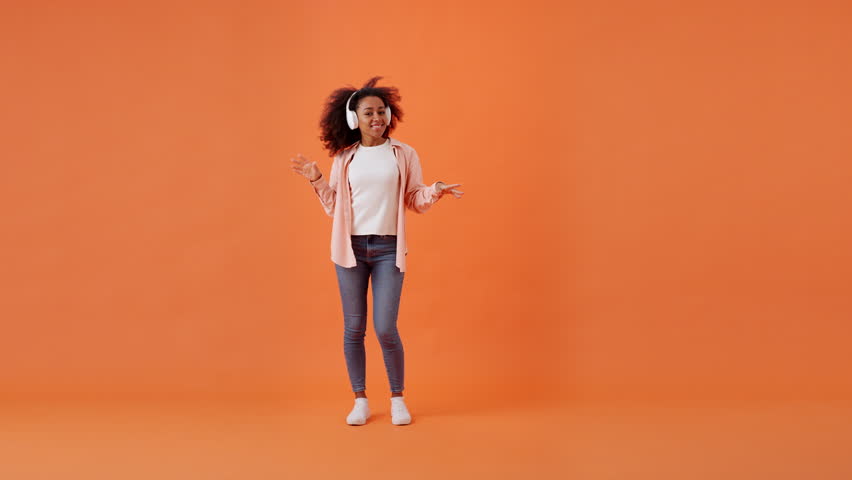 Happy African American woman dances listening to music on headphones. Lady relishing active lifestyle and engaging in dynamic body movements in orange premise Royalty-Free Stock Footage #3400577779