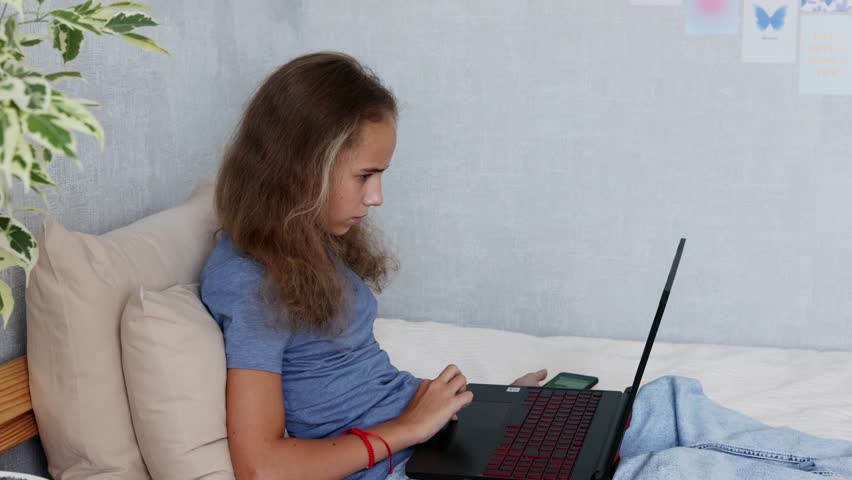 A cute blonde teenage girl is typing on a laptop while sitting on her bed in the bedroom