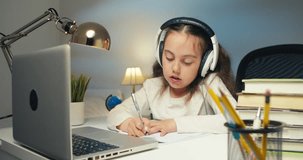 Little schoolgirl in headphones using laptop, studying online at home, interested happy student typing on keyboard, looking at computer screen, watching webinar, online course, doing homework