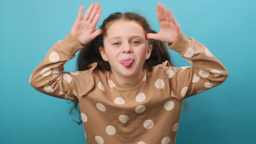 Portrait of funny little girl kid sticking out tongue and grimacing looking at camera, holding hands near temples, posing isolated over plain blue color background wall in studio. Misbehavior concept Royalty-Free Stock Footage #3400733223