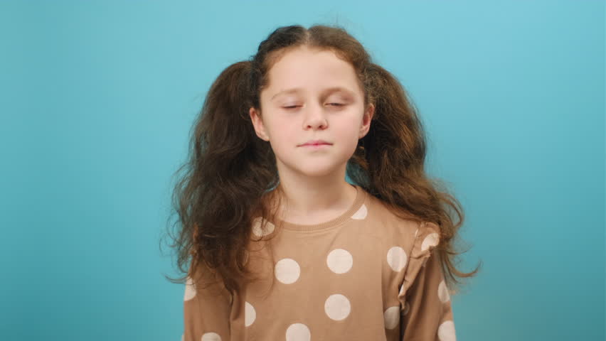 Portrait of cute peaceful preteen girl praying to God, making prayer gesture, looking at camera, posing isolated over plain blue color background wall in studio. Sincere kids faith. Religion concept