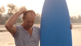 Mature male surfer with blue surfboard ready to catch waves, cleaning and washing face and hair from sand and water after swimming in tropical area. Learning swim, surfing on vacation, resort. Video