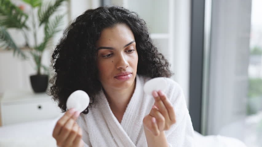Close-up portrait of positive curly haired multinational woman in bathrobe with clean smooth flawless skin, removing make-up with cotton pads, spa, therapy, treatment, on background of bright bedroom.