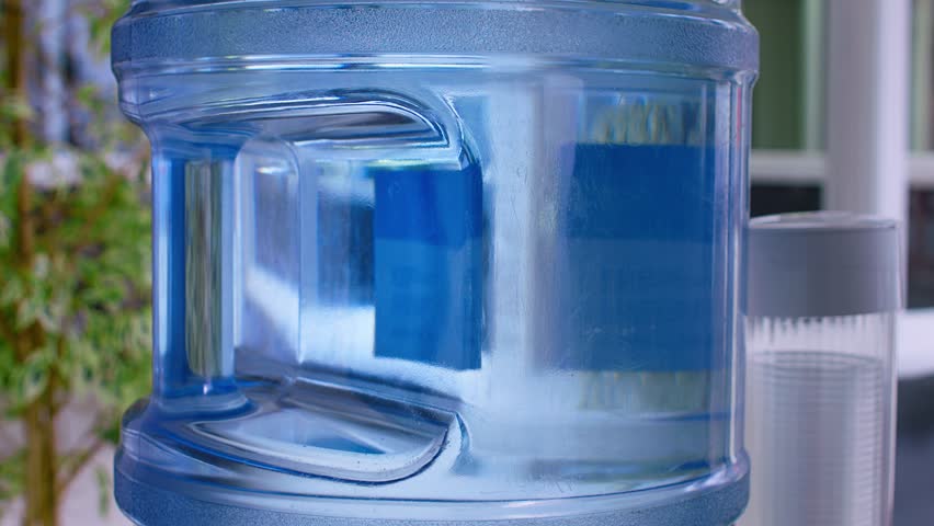 Large bubbles rise through the plastic tank of an office water cooler. The cooler has paper cups mounted to the side and office workers move around in the background of the shot.