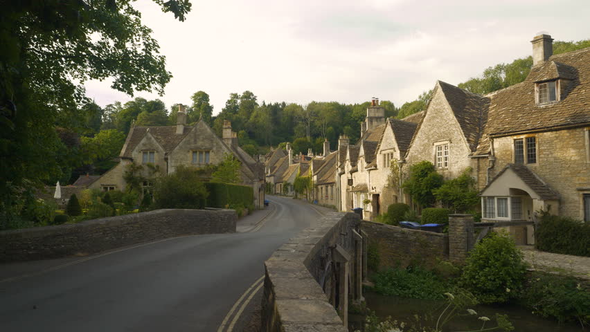 Crossing bridge towards idyllic countryside village with charming stone houses. Picturesque terraced cottages placed along a winding asphalt road below a green hill in delightfully pretty Castle Combe
