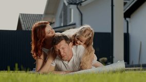 American dream family is resting outdoors near the house, lying on the grass and watching funny video in tablet. Parents kiss daughter. Little girl in joyful emotions smiles and looks at the camera