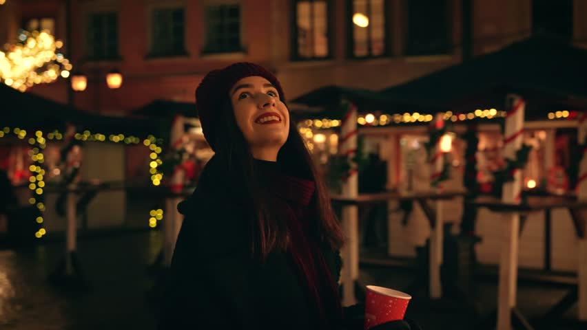 A cheerful girl drinking a hot drink at the Christmas market near the garlands, enjoying the festive atmosphere. Atmosphere of Christmas and New Year.