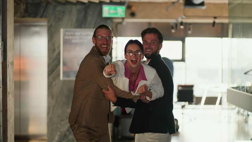 A blond man in a brown suit and a brunette man in a blue jacket carry in their arms a brunette girl in a pink shirt and white jacket. Playing in the office. The girl extended her hand forward and was