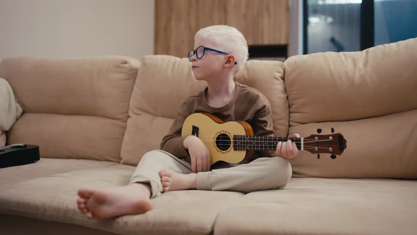 Little albino boy with white hair color in round blue glasses plays the ukulele while sitting on a cream sofa in a modern apartment in the evening. A little albino boy with white hair in blue glasses