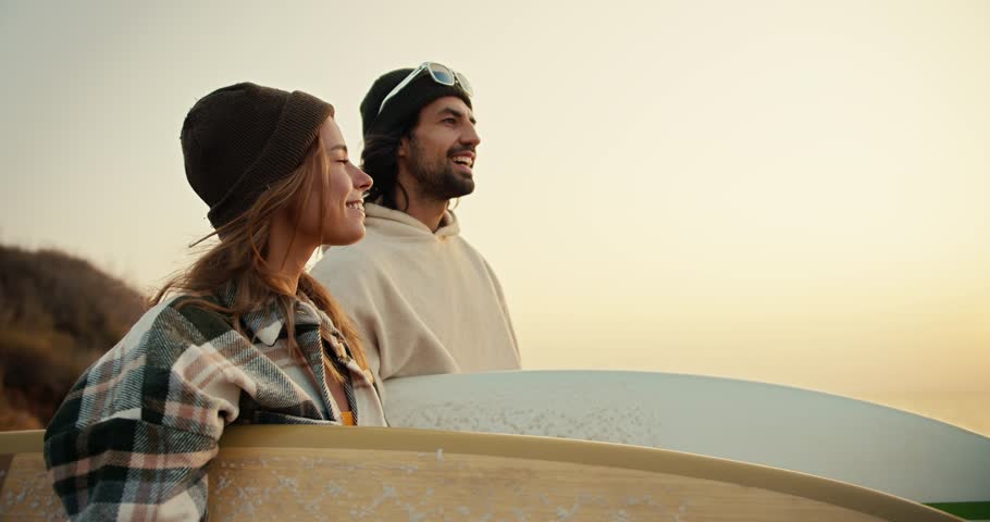 A brunette man in a black hat and white top points somewhere in the direction of his blonde girlfriend in a plaid shirt while they stand on the seashore at dawn in the morning and hold surfboards in Royalty-Free Stock Footage #3401102855