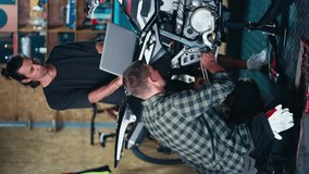 Vertical video: 2 mechanics repairing a bike. A brunette man in a gray T-shirt looks at the screen of a gray laptop and a man in a checkered shirt repairs a motorcycle in a workshop garage - Powered by Shutterstock - Get 15% off with code: PIKWIZARD15