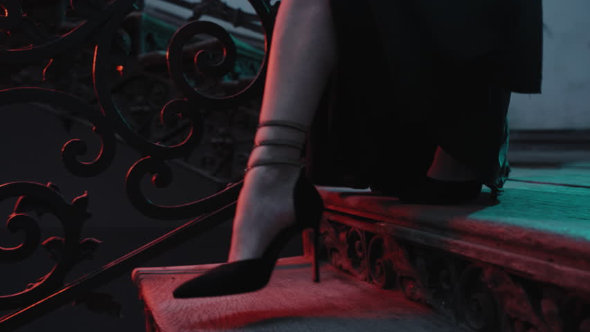 In a close-up, a woman in high heels walks down the fancy stairs, looking stylish in a black dress. It