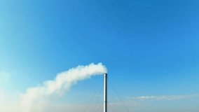 Thick plumes of white smoke billow from the towering industrial plant, shrouding the area in a hazy veil, a stark contrast against the clear blue sky. Air quality and global warming concept. Drone.
