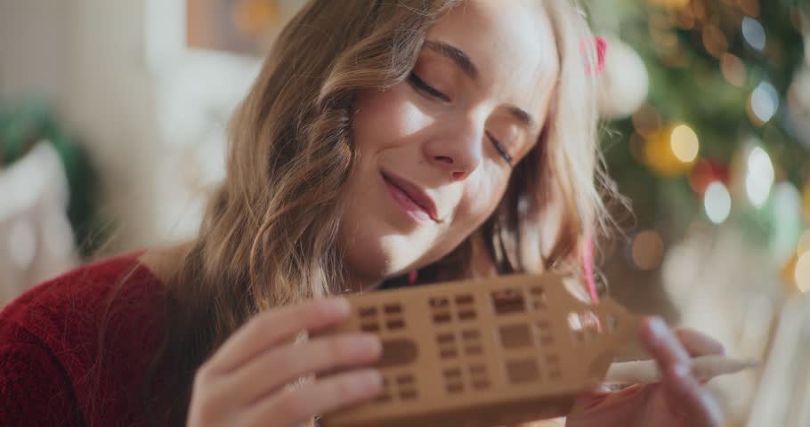 Beautiful young woman using white sketch pen to color cardboard house ornament during Christmas at home