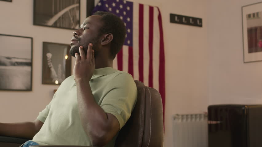 Portrait of young cheerful African American man sitting against USA flag in barbershop and posing for camera with smile
