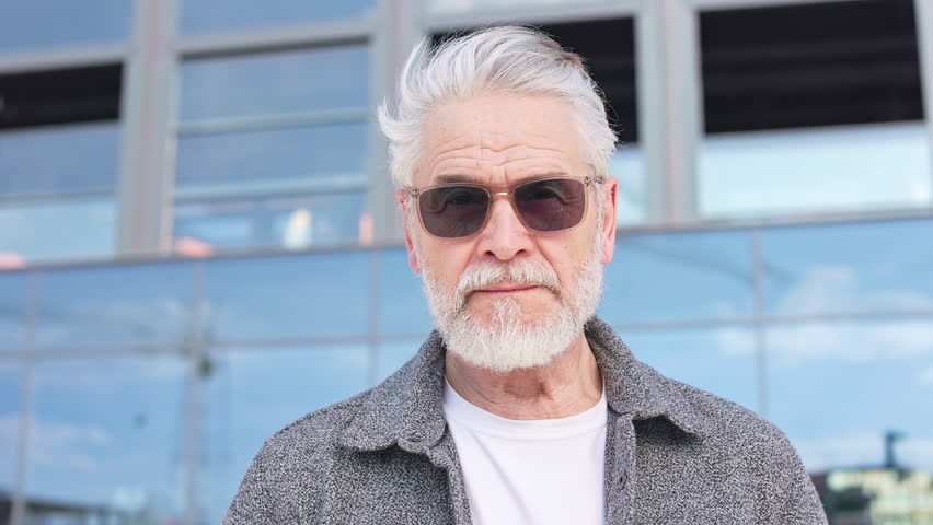 Portrait of concentrared aged man looking at camera and not moving while standing near contemporary mirror building facade. Calm pensioner in sunglasses thinking about life during free time outside. Royalty-Free Stock Footage #3401441349