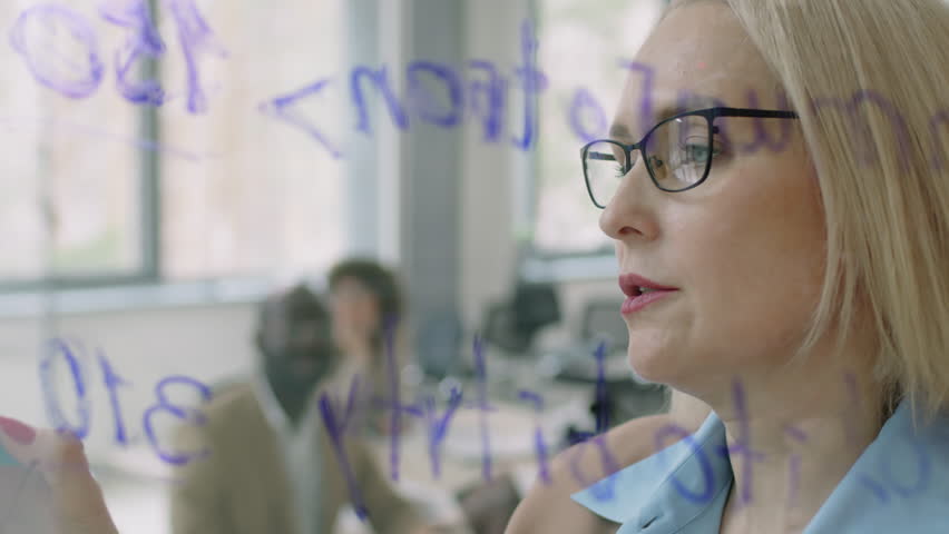 Shot through glass of Caucasian female teacher standing in well-lit meeting room holding special seminar while writing on glass wall Royalty-Free Stock Footage #3401443225