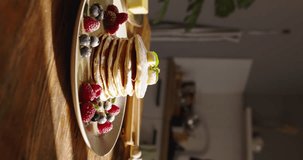 Pancakes in kitchen, aesthetic meticulous arrangement of berries, powdered sugar on plate. Breakfast aesthetics, enjoying delightful meal, wooden table under the warm morning sunlight. Vertical video