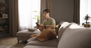 Attractive lady reclining contentedly on sofa, basking in sunshine, savoring free time with boyfriend, communicating with him online on smartphone. Psychological balance, warmth of sun. Cinematic AD