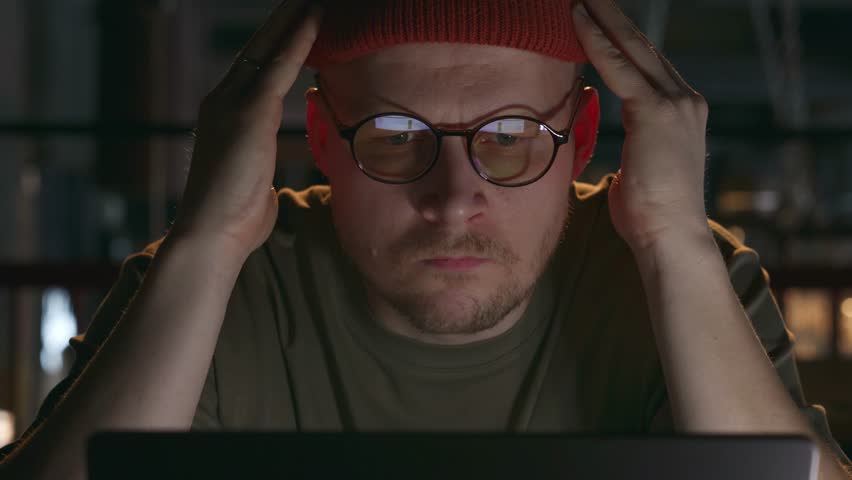 A close-up portrait of a millennial hipster man who works at a laptop and looks at the computer screen in a puzzled way and does not know how to solve the problem. A difficult or hopeless situation Royalty-Free Stock Footage #3401590507