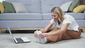 Mature woman doing gymnastics workout watching online video course on laptop sitting on floor in living room at home. Beautiful female doing stretching exercises while listening to instructor call