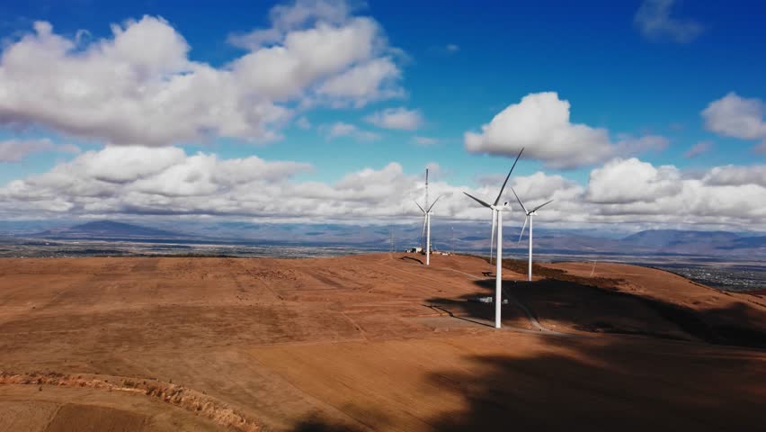 Dry agriculture fields and wind generators standing there with mountain range on background filmed by drone at autumn day under blue sky. Integration of renewable energy technology into farm landscape