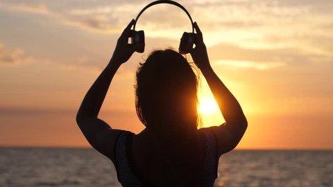Silhouette of young woman wearing headphones have fun listening music on the beach at amazing sunset in slow motion. 1920x1080
