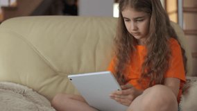 Child Browsing Social Media. Portrait of Cute Girl Playing Game on Tablet PC Relaxing in Living Room. Child Typing Using Tablet PC Sitting on Sofa at Home, Homeschooling, Distant Remote Education. 