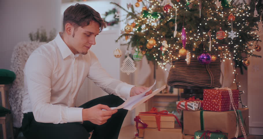 Happy young man reading letter while crouching by Christmas presents and decorated tree at home