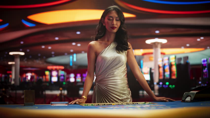 Portrait of a Professional Female Croupier Looking at the Camera and Welcoming You to a Table. Beautiful Asian Dealer in Silver Dress Dealing Playing Cards in a Casino for a Game of Baccarat