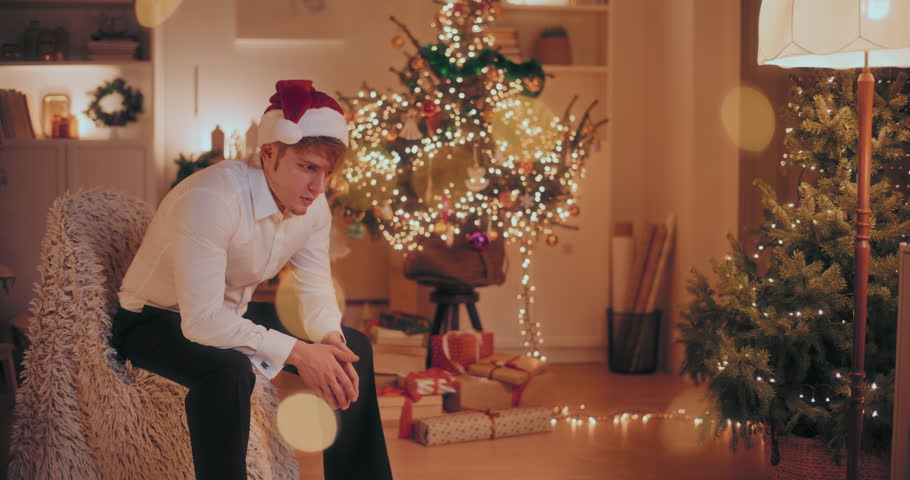 Sad young man in formals and Santa hat sitting on chair at illuminated home during Christmas