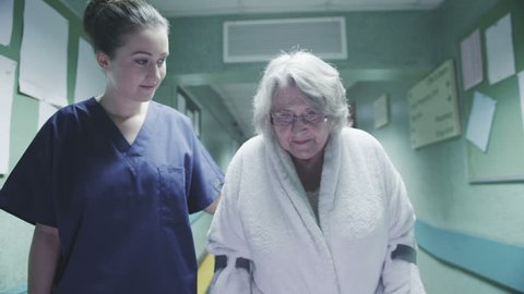 An attractive female medical assistant or nurse helps an elderly lady on crutches to take a walk down a busy hospital corridor.