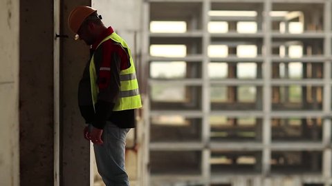 Worker knocks his head against the wall. Stressed builder in hard hat stands on construction site and knocks his head against the concrete wall, self-condemnation and self-torture