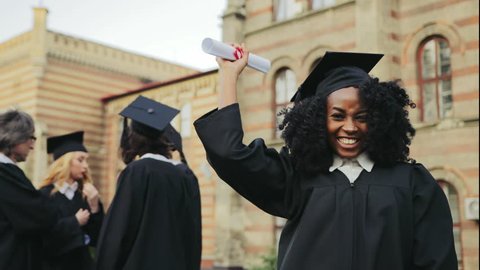 Portrait of the African American happy young graduated woman posing to the camera, showing her diploma and making YES gesture in front of the University. Graduates with professor on the background