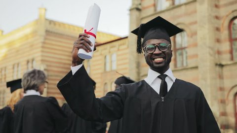 Portrait of the African American happy young graduated man posing to the camera and showing his diploma in front of the University. Graduates with professor on the background. Outdoors