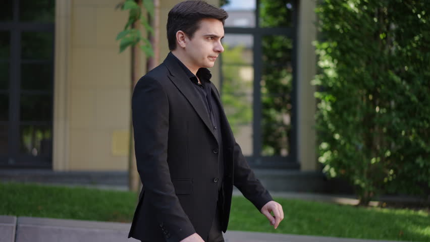 A young man in a black formal suit turns away, covers his face with his hands and sneezes while standing on the street in a city park