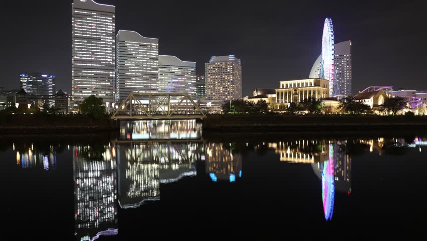 TIMELAPSE: Yokohama skyline with iconic giant ferris wheel and skyscrapers of the illuminated waterfront in the bay, reflect in the water at night. Cityscape of Yokohama, Japan.