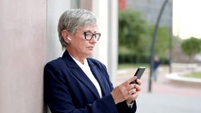 Horizontal video of mature businesswoman leaning on a wall using phone and smiling looking at camera