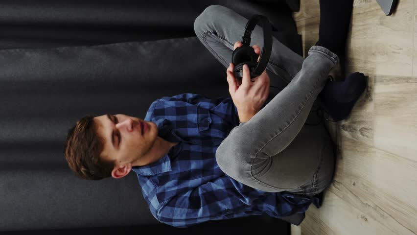 Young man listening to music in headphones and singing. Male puts on headphones and starts singing while sitting on floor in modern living room. Lifestyle concept. Music concept. Vertical video