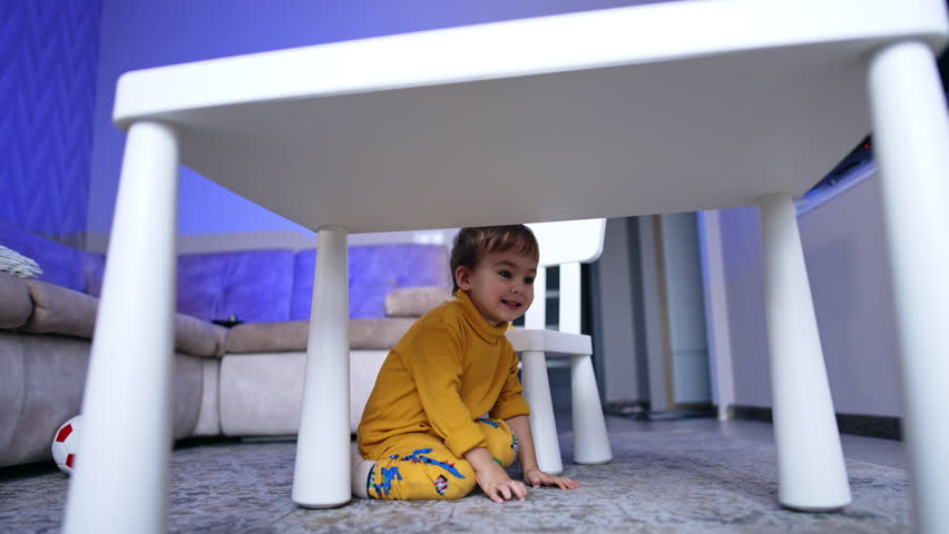 Cute two year old toddler sitting on his knees on the floor near the desk. Baby boy in yellow clothes crawls by the floor.