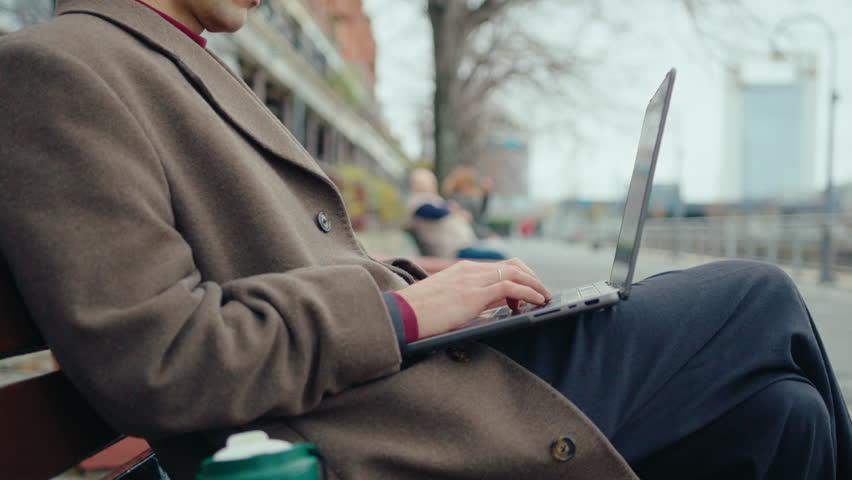 Businessman in elegant coat sitting on bench on the street and typing on laptop, working online outdoors in the city. Close-up shot
