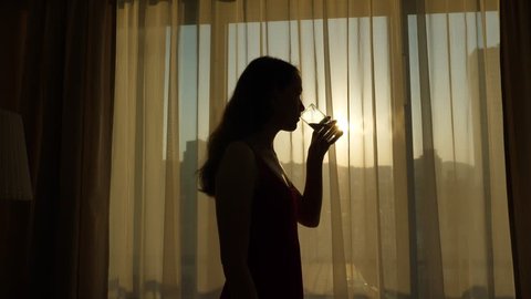 Young woman take one gulp of water, silhouetted view against living room window, sunrise outside. Half length shot of long haired lady, enjoying good morning time, healthy lifestyle concept